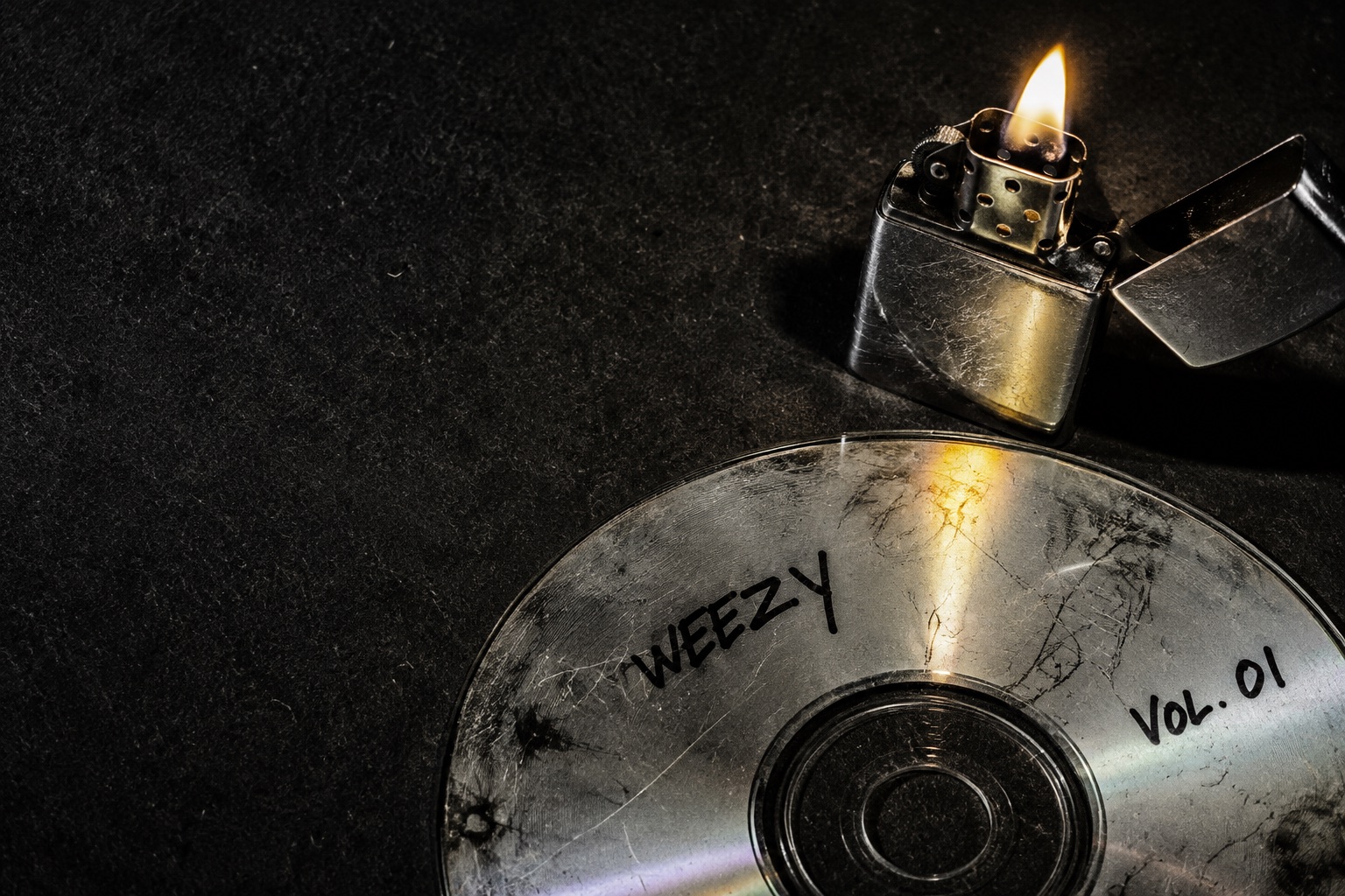 A scratched Weezy CD beside a lit chrome lighter on a dark table